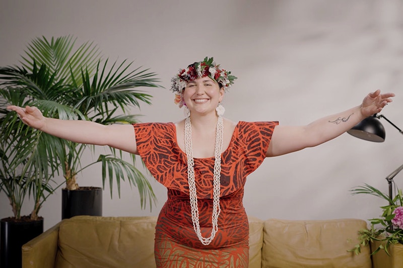 A woman is partway through dancing with her arms spread out and she is smiling at the camera. She is wearing a floral wreath on her head.