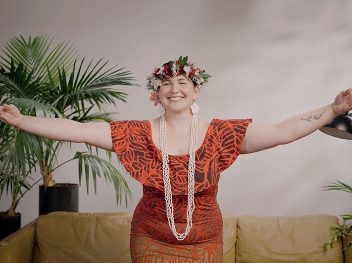 Still from Kate Ngatokorua Not Your Dusky Maiden A woman is partway through dancing with her arms spread out and she is smiling at the camera. She is wearing a floral wreath on her head.