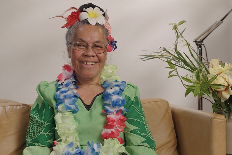 A woman wearing flowers in her hair and a flower lei is sitting on a sofa smiling off camera.