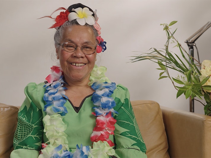 Still from Not your dusky maidens: Mata Atonio A woman wearing flowers in her hair and a flower lei is sitting on a sofa smiling off camera.