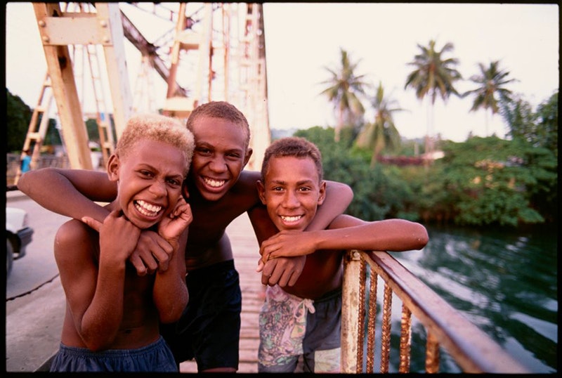 Three young boys are standing on a bridge with arms around each other's shoulders. There are palm trees in the background.