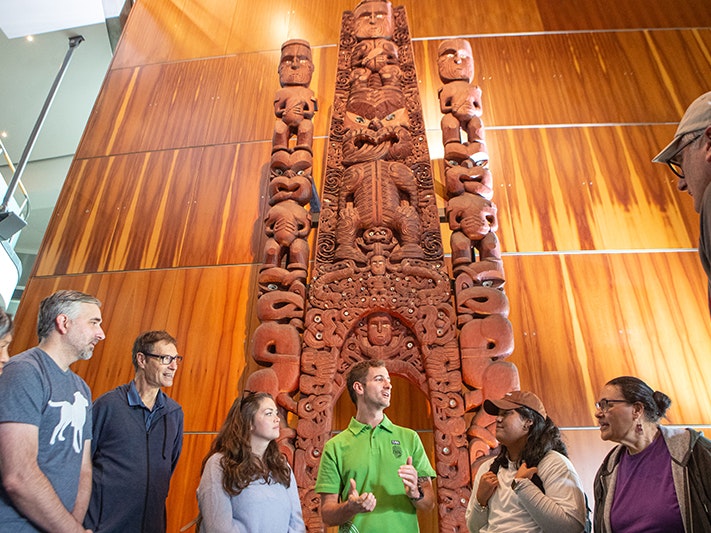 Te Papa tours, 2025. Photo by Jeff McEwan. Te Papa (267134) Several people are gathered at the base of a large waharoa carving in a museum. They are listening to the person in the middle.