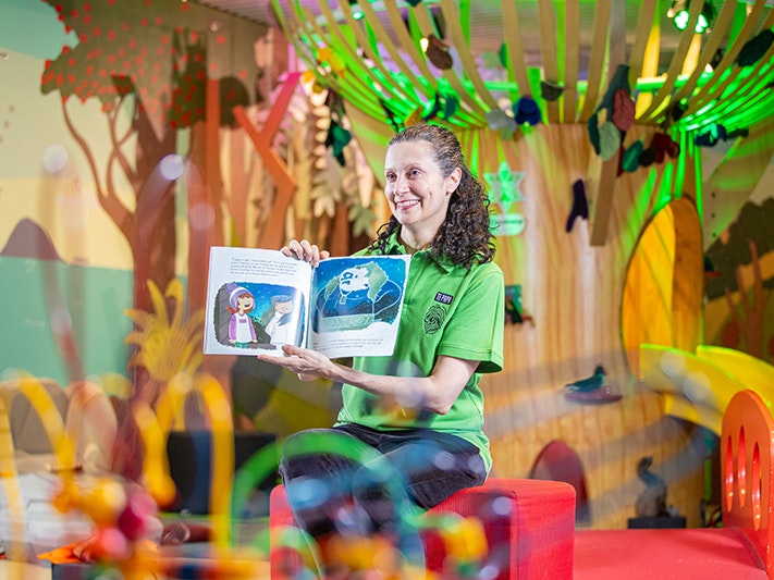 StoryPlace at Te Papa. Photo by Jeff McEwan. Te Papa (270458) A woman in a green shirt is holding a picture flat children's book up and reading to children off camera.