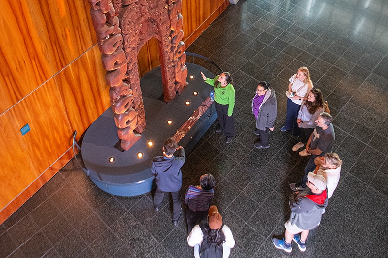 A person in a green shirt is pointing at a wooden carving and talking to people standing around him.