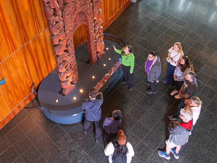 Te Papa Tours. Photo by Jeff McEwan. Te Papa (267138) A person in a green shirt is pointing at a wooden carving and talking to people standing around him.