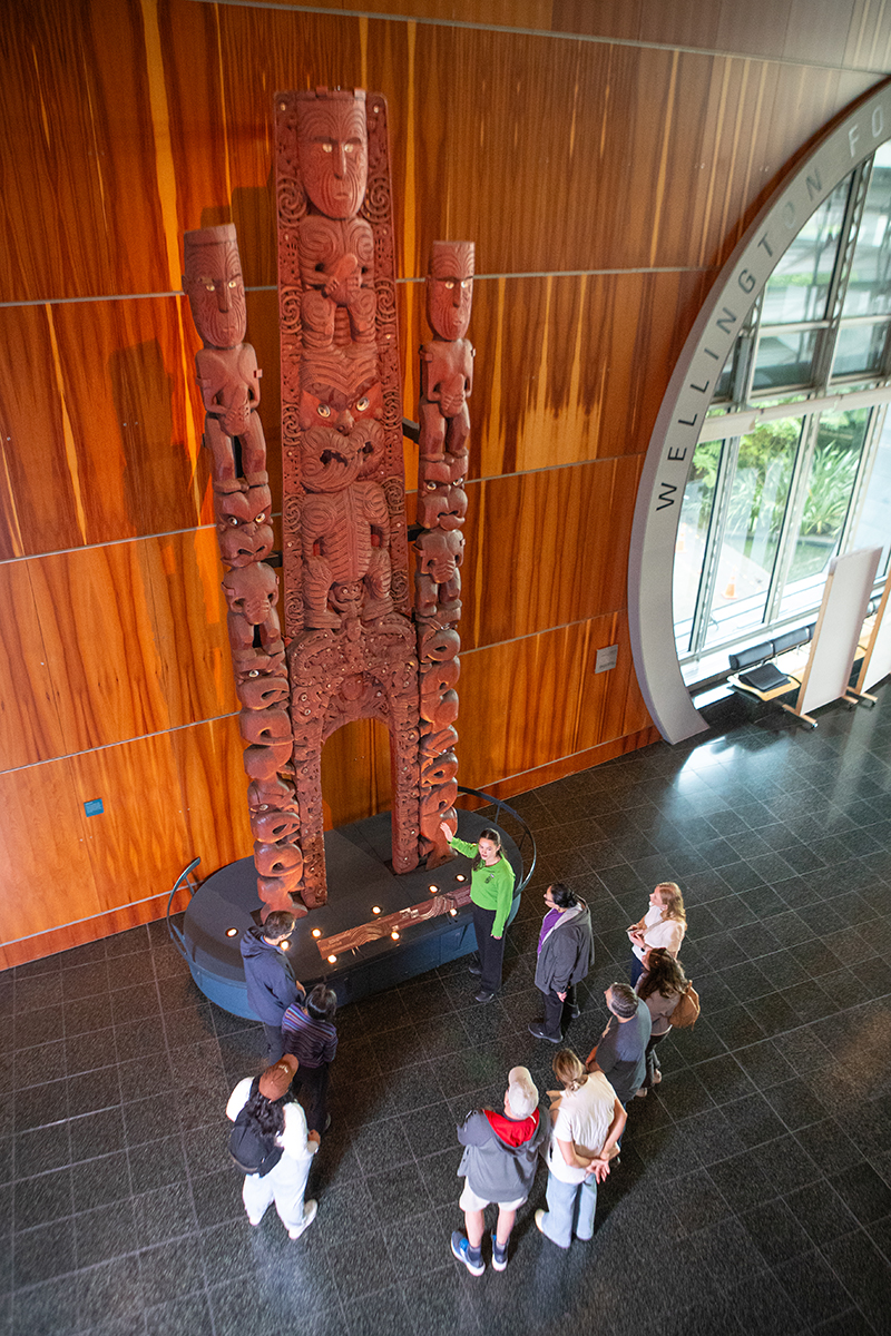 A man in a red top is pointing at a very tall wooden carving. He is describing something to the man crouched next to two small children.