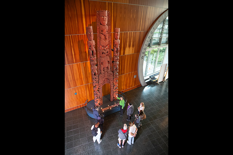 A man in a green top is pointing at a very tall wooden carving. He is describing something to the man crouched next to two small children.