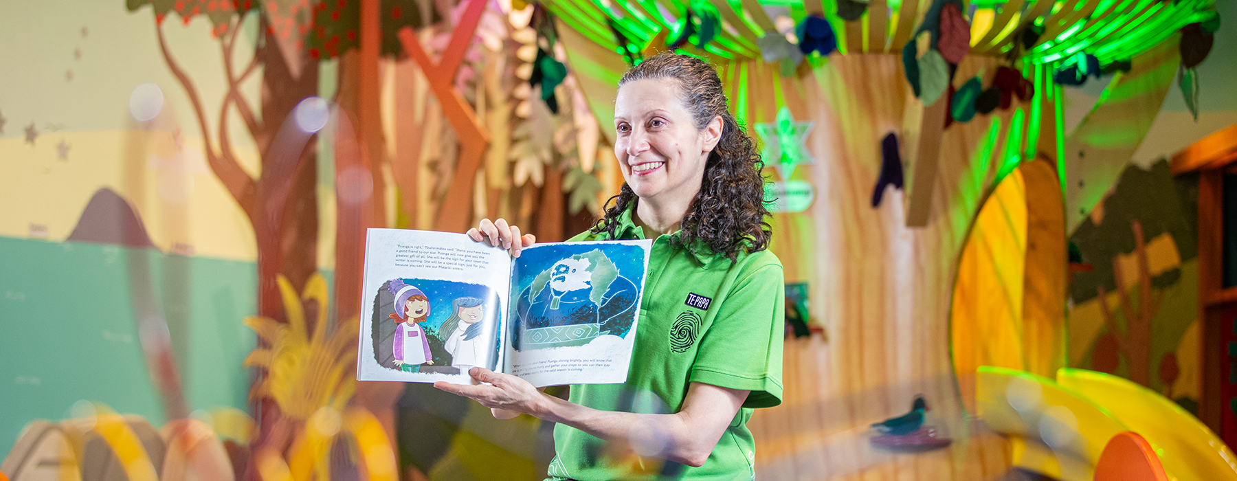 A woman is holding up a picture flat children's book and reading it to children out of shot.