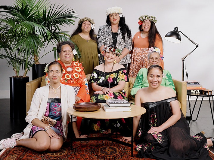 Still from We are not your dusky maiden. Te Papa eight women in bright clothing are seated or gathered around a sofa and smiling at the camera.