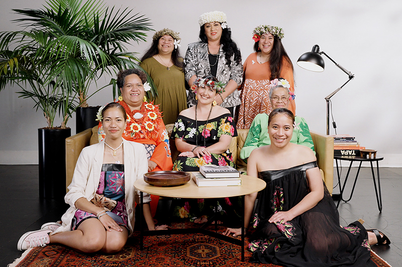 eight women in bright clothing are seated or gathered around a sofa and smiling at the camera.
