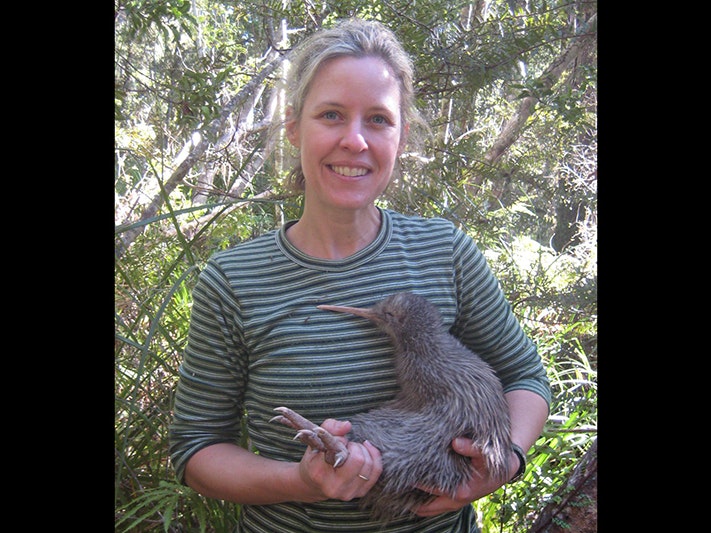 Kiwi researcher Kristina Ramstad holding a rowi. Photo by Rachael Abbott A woman in a striped top is standing in the forest and holding a live kiwi bird.