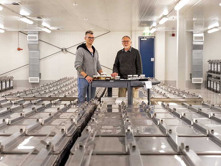 Curators Thom Linley (L) and Andrew Stewart (R) with specimens to be housed in the Te Papa Biodiversity Research Centre. Te Papa 2025. Two people are standing in the middle of a room with a trolley of fish specimens in jars in front of them. Surrounding them are large metal tanks full of fish you cannot see.