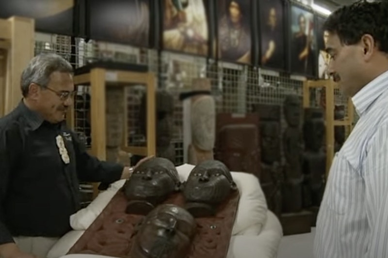 Two men are standing either side of a carving that is laid on a pillow. There is a museum store space in the background.