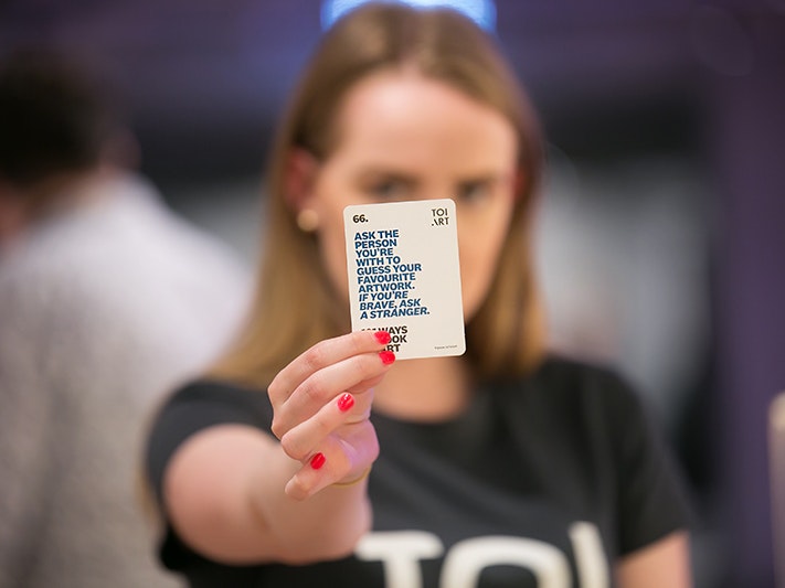 Toi Art Opening Weekend Events and Programmes. Photo by Jo Moore. Te Papa (103299) A woman in a black tshirt is holding up a small card that says "Ask the person you're with to guess your favourite artwork. If you're brave, ask a stranger."