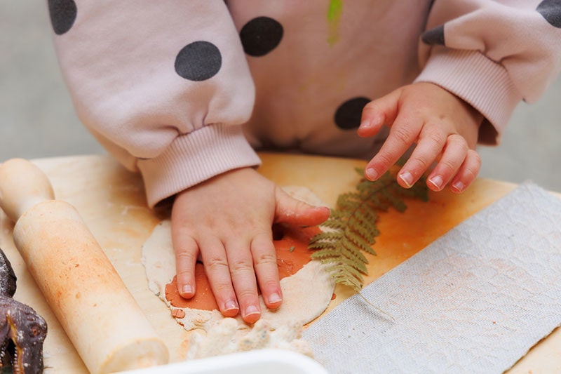 A child's hands press down a piece of clay next to a leaf and rolling pin.