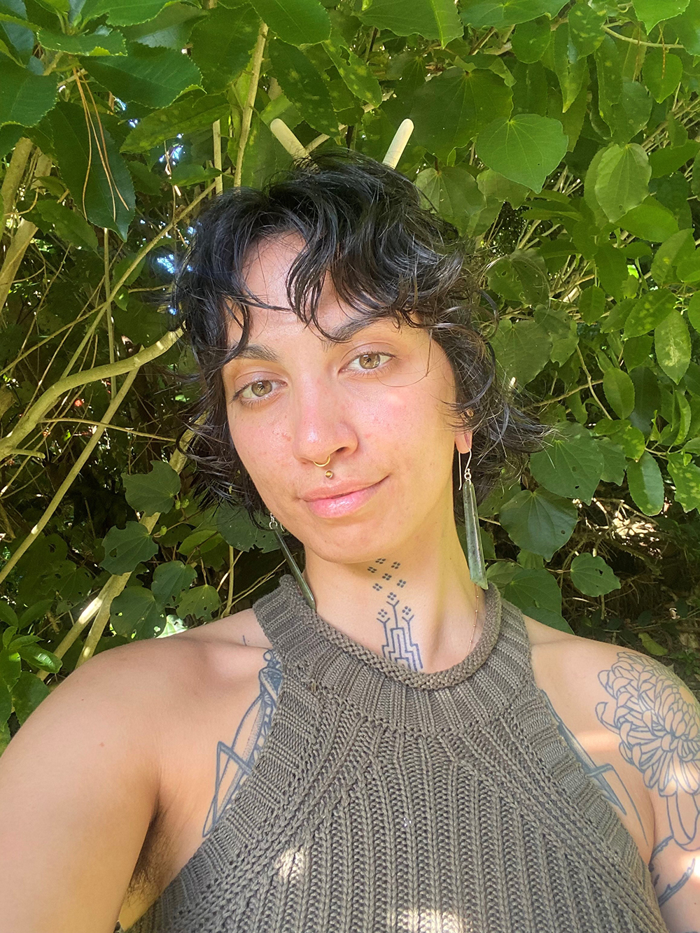 A person with a bare-shouldered top and tattoos is standing in front of kawakawa leaves.