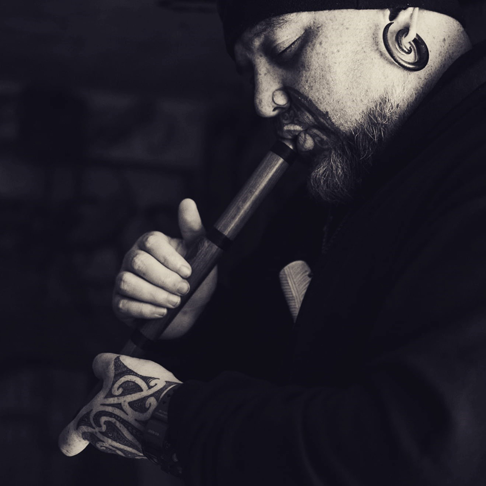 A man with a beard and a tā moko is blowing into a wooden Māori instrument.