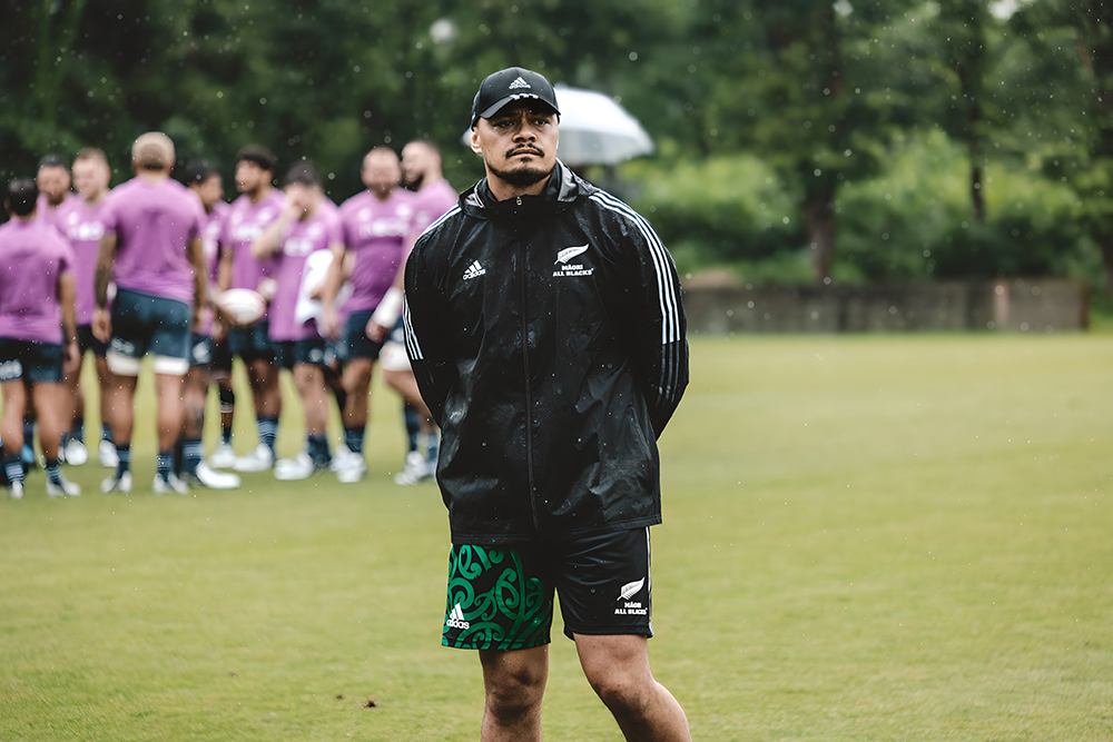 A man is in a cap and shorts standing on a filed in the rain. There is a team of rugby players in a purple uniform behind him.