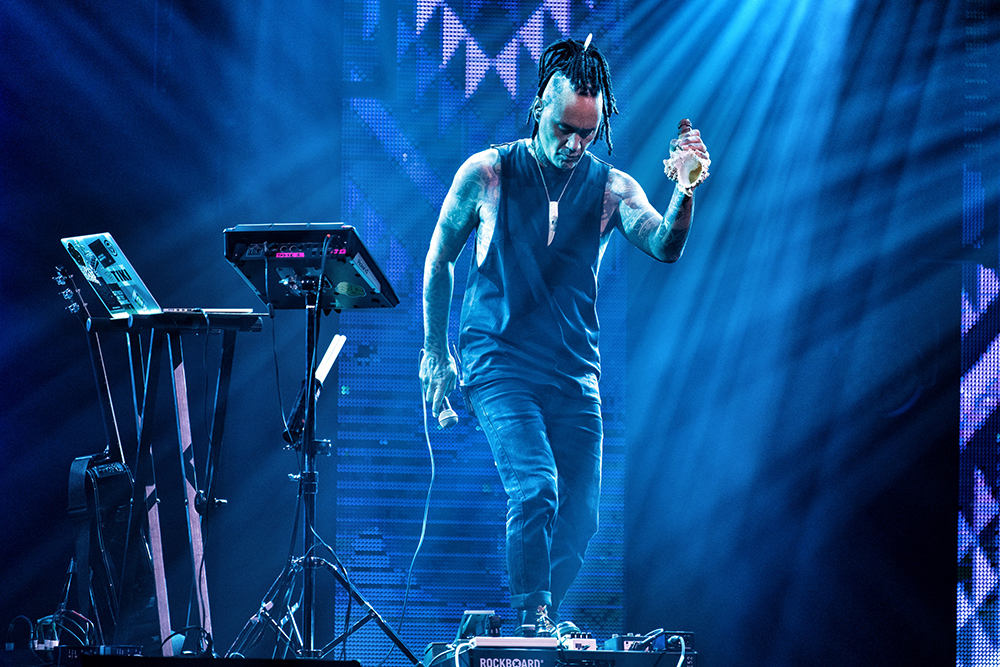 A man with dreadlocks pulled back in a ponytail is wearing a singlet and jeans and is stepping on a loop pedal on a stage. There are other instruments beside him.