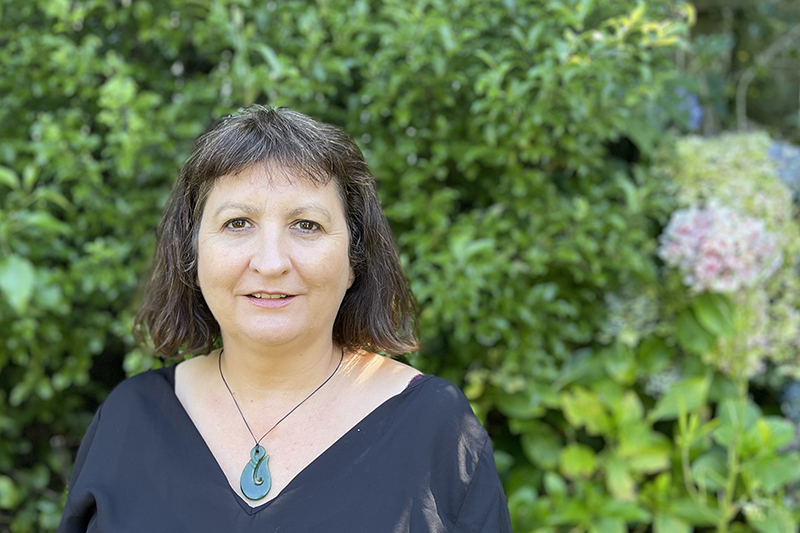 A head and shoulders photo of a woman standing in front of green bush.