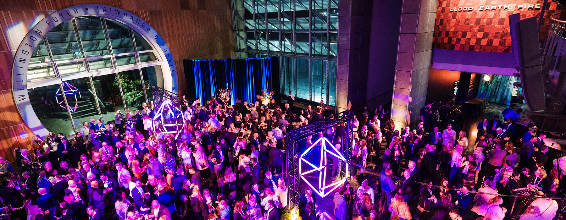 Many people gathered in a large museum foyer at night. There is a purple lighting hue across them all.