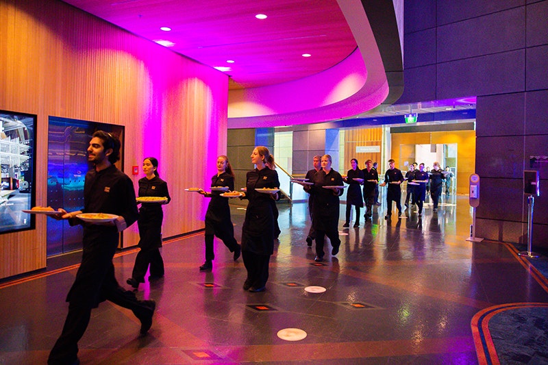 Several people dressed in black are carrying platters of food through an open foyer in a large building.