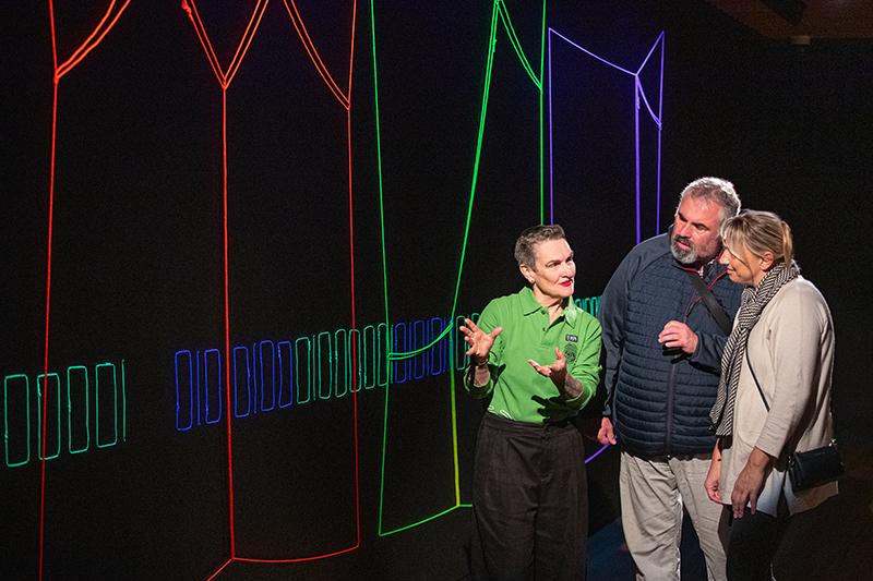 A person is talking to two other people about an artwork on the wall that is brightly coloured string on a black wall.