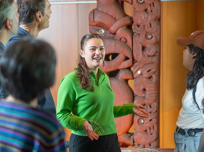 Te Papa Tours, 2025. Photo by Jeff McEwan. Te Papa (267131) A woman in a green shirt is talking to a group of people while standing in front of a Māori carving.