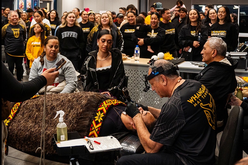 A person wearing a headband torch is tattooing the chin of a woman lying on a table under a feather cloak. They are surrounded by a lot of people.
