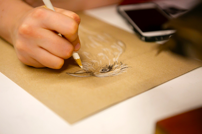 A close up of a hand drawing a bird on a piece of brown paper.