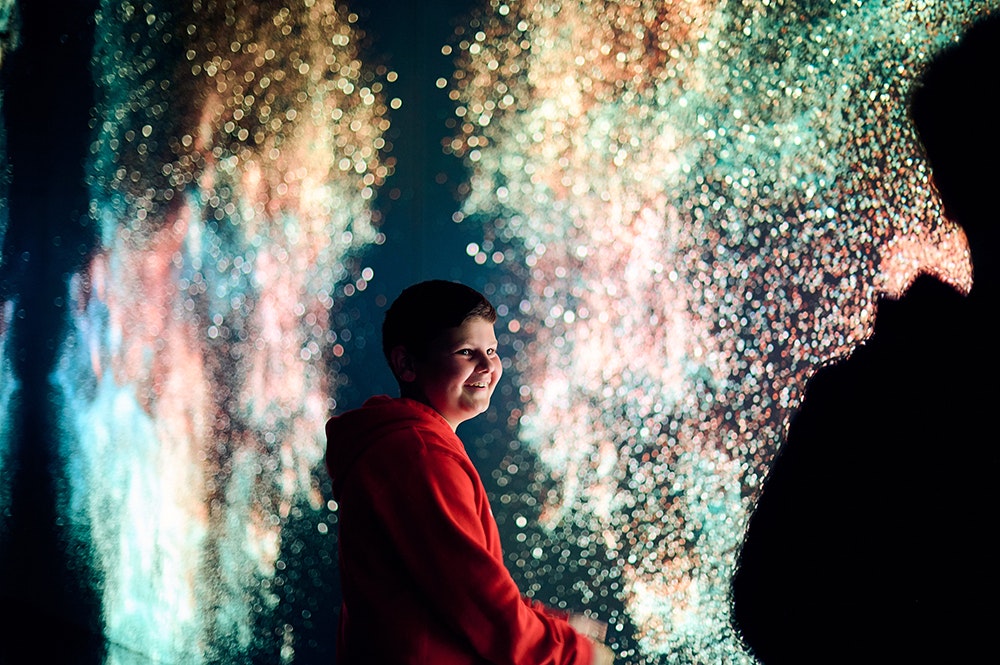 A child is smiling up at someone with them. They are both in front of a gigantic screen with dots of light and swirling patterns.