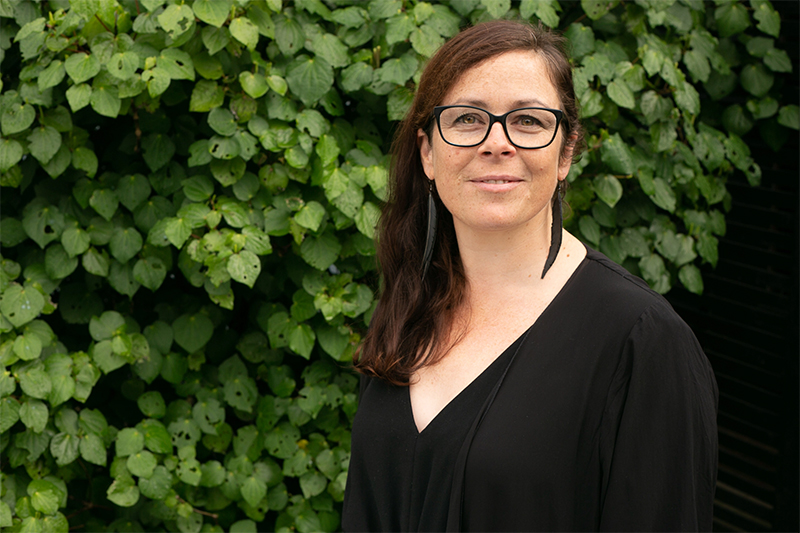 Head and shoulders photo of a woman with shoulder-length dark hair, glasses and a dark top. She is standing in front of a kawakawa bush.
