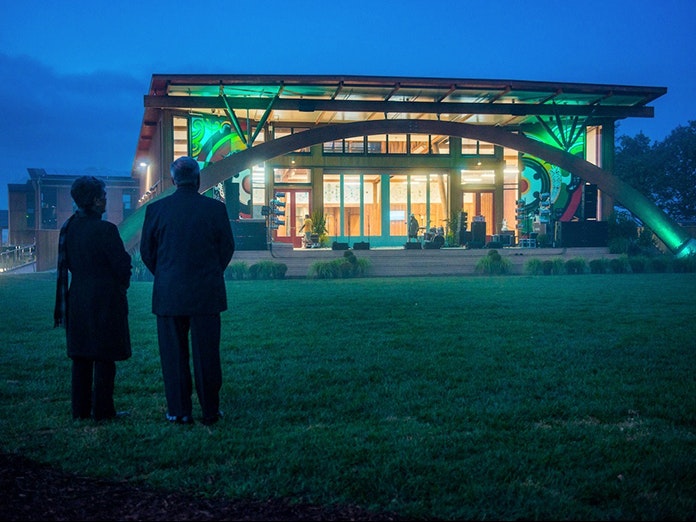 Still from Ever the Land. Courtesy of Sarah Grohnert  Two people with their backs to the camera are looking at a lit-up building in a grass field at twighlight.