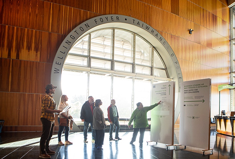 A person in a green top is pointing to a sign in front of a large circular window. There is a group of people looking on.