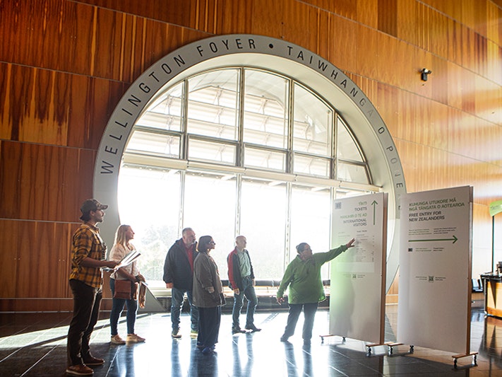 Te Papa Tours, 2025. Photo by Jeff McEwan. Te Papa (267120) A person in a green top is pointing to a sign in front of a large circular window. There is a group of people looking on.