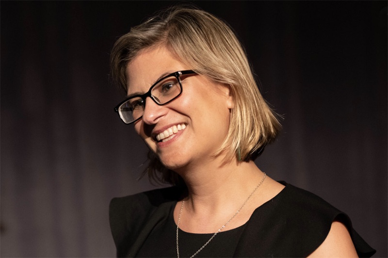 A head and shoulders photo of a woman with blonde hair and glasses smiling at someone off screen.