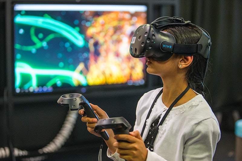 A young child is wearing a Virtual Reality headset and holding two handsets.