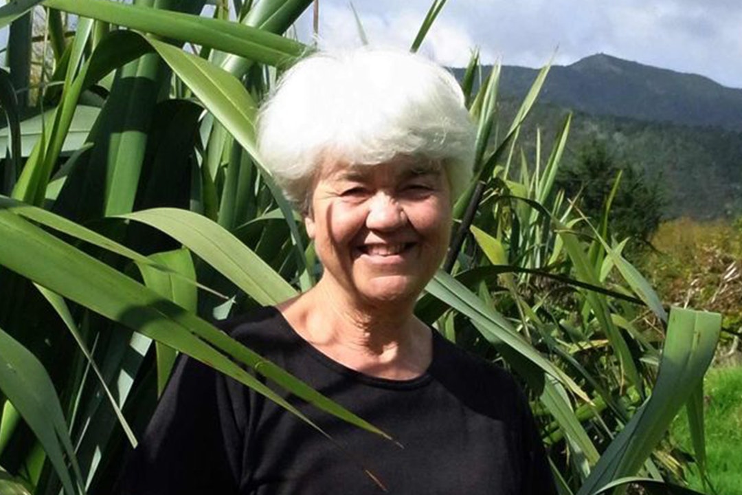 A woman with white hair is standing in front of a large flax bush on a sunny day.