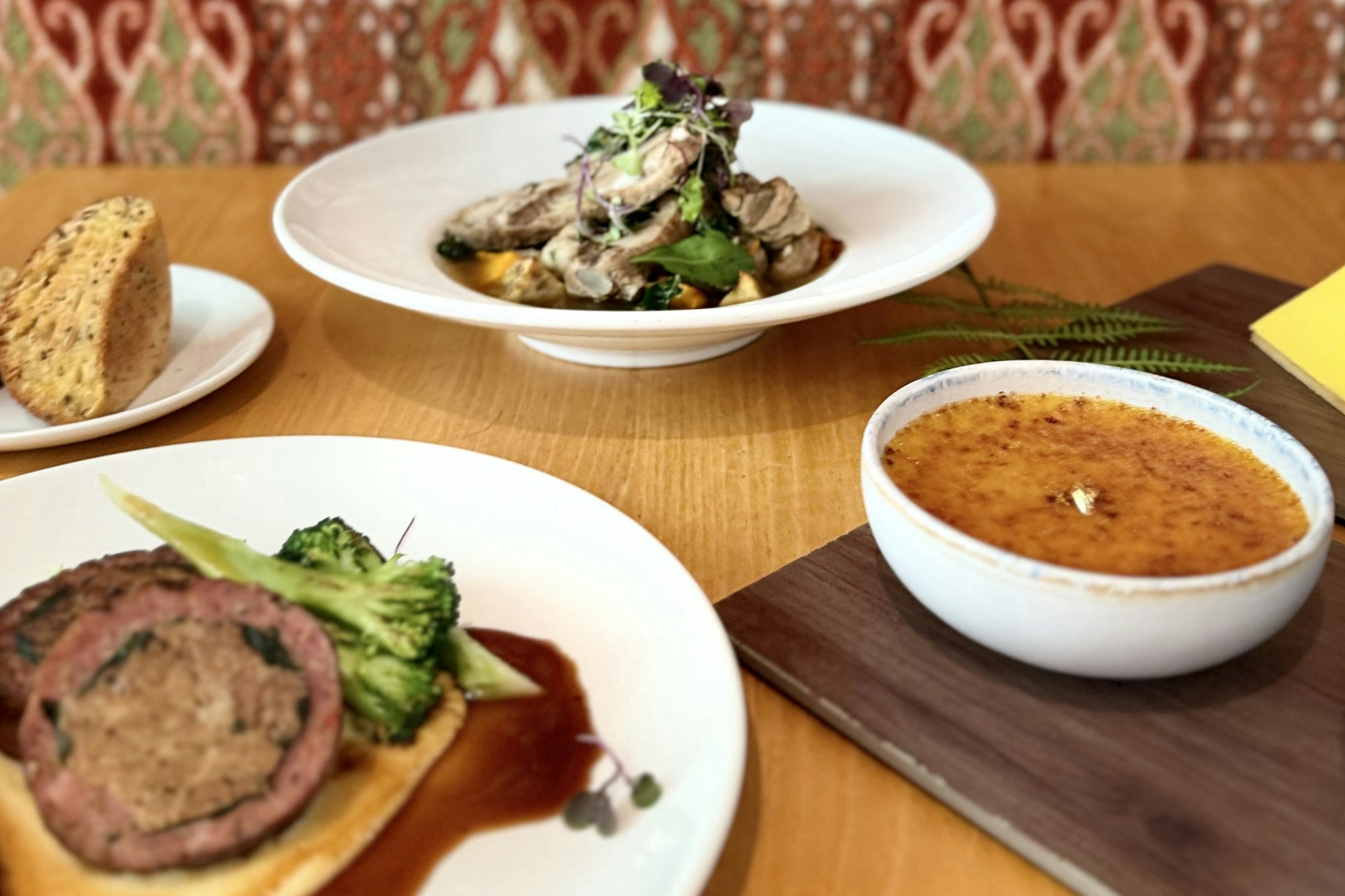 Plates of food on a table. Two are savoury dishes, one is sliced bread, and one is a bowl of dessert.
