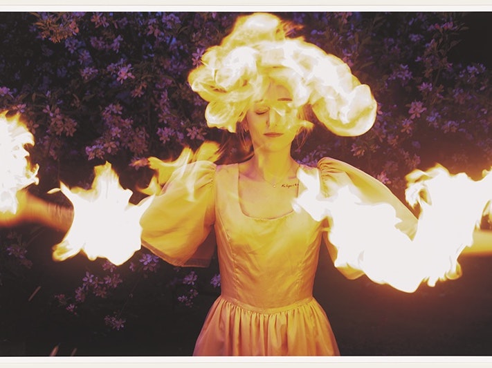 Tia Ranginui, The tangi of Rūaumoko, 2023. Purchased 2024. Te Papa (O.052215) A woman wearing a silk gown with puffy sleeves stands with arms outstretched in front of a background of purple flowers. Flames engulf her head and arms while she smiles serenely with eyes closed.