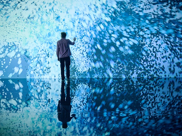 Marshmallow Laser Feast: Works of Nature, 2023, ACMI. Photo © Eugene Hyland. Man standing in front of immersive installation of swirling blue light witha hand raised to touch it.