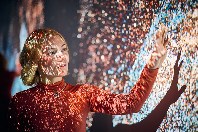 A woman has a lightshow playing over her face and she has an arm up near the screen it is projecting on.