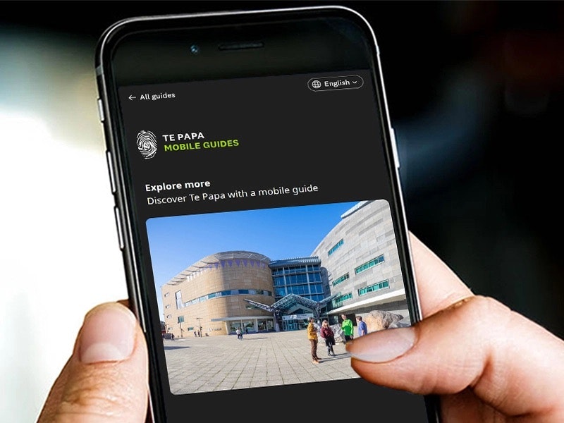 A close up of a hand holding a mobile phone that has an image of Te Papa from the front forecourt.
