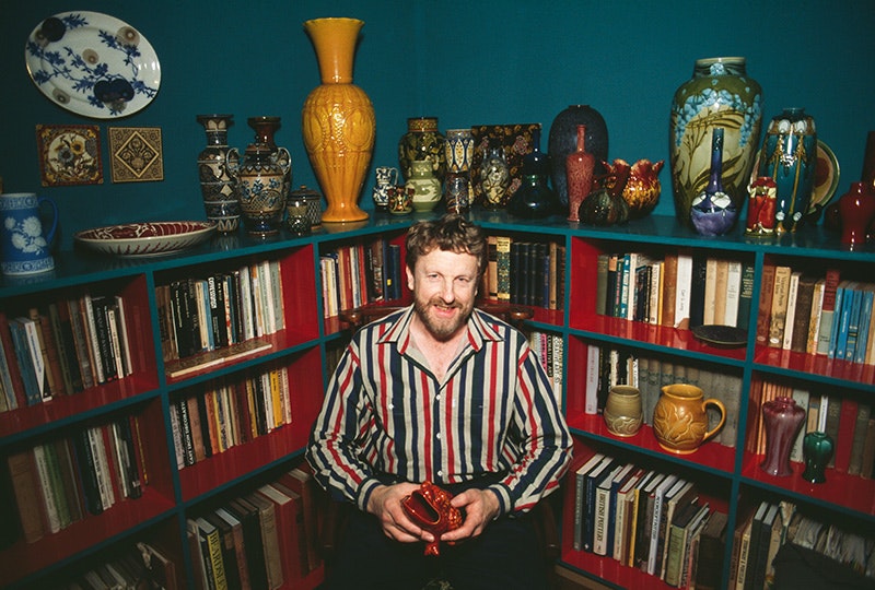 A man in a stripey shirt and beard is smiling and holding a pot in front of a collection of books and pots.
