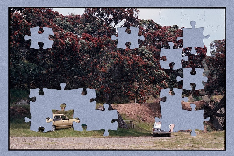 Two cars are parked under some large trees that have red flowers on them. Next to each car is a camping tent. There is someone sitting in front of one of the tents in a fold-out chair. The image has jigsaw piece shapes missing.