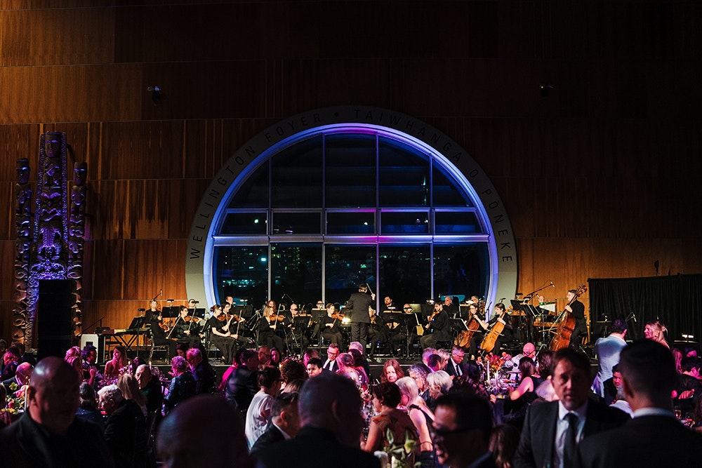 A large room with a round window is full of people at a party. The room is light in dark purples and blues.