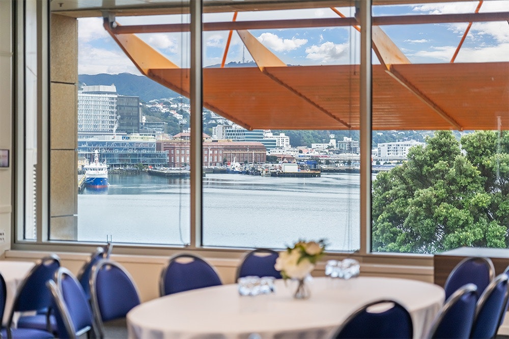 A white-clothed table with blue chairs around it next to a window with a view of Wellington Harbour.