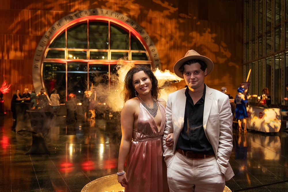 Two people are standing together in the foreground and smiling at the camera. There is an event with dry ice in going on in the background.