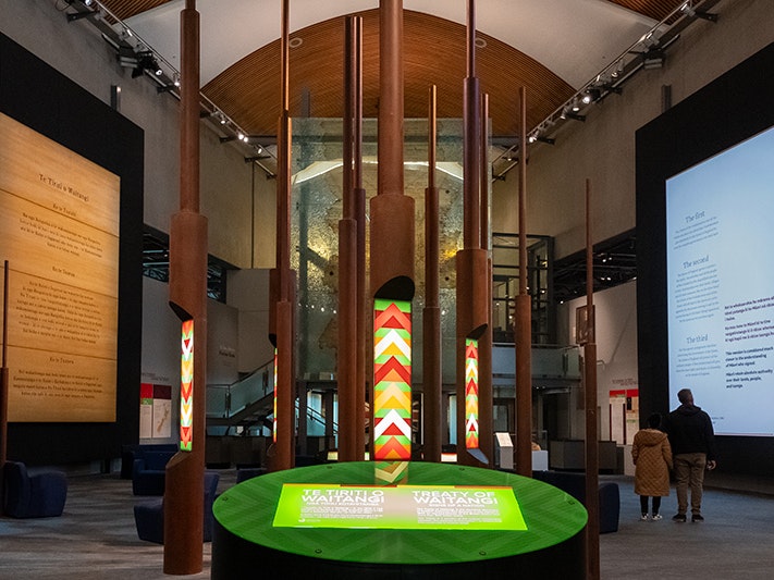 Te Tiriti o Waitangi: Ngā tohu kotahitanga, 2024. Photo by Jane Harris. Te Papa (244624) A wide view of the Tiriti exhibition space in Te Papa, with the sgreen signage in the foreground, the tall pou in the mid-ground and the two walls of writing either side.