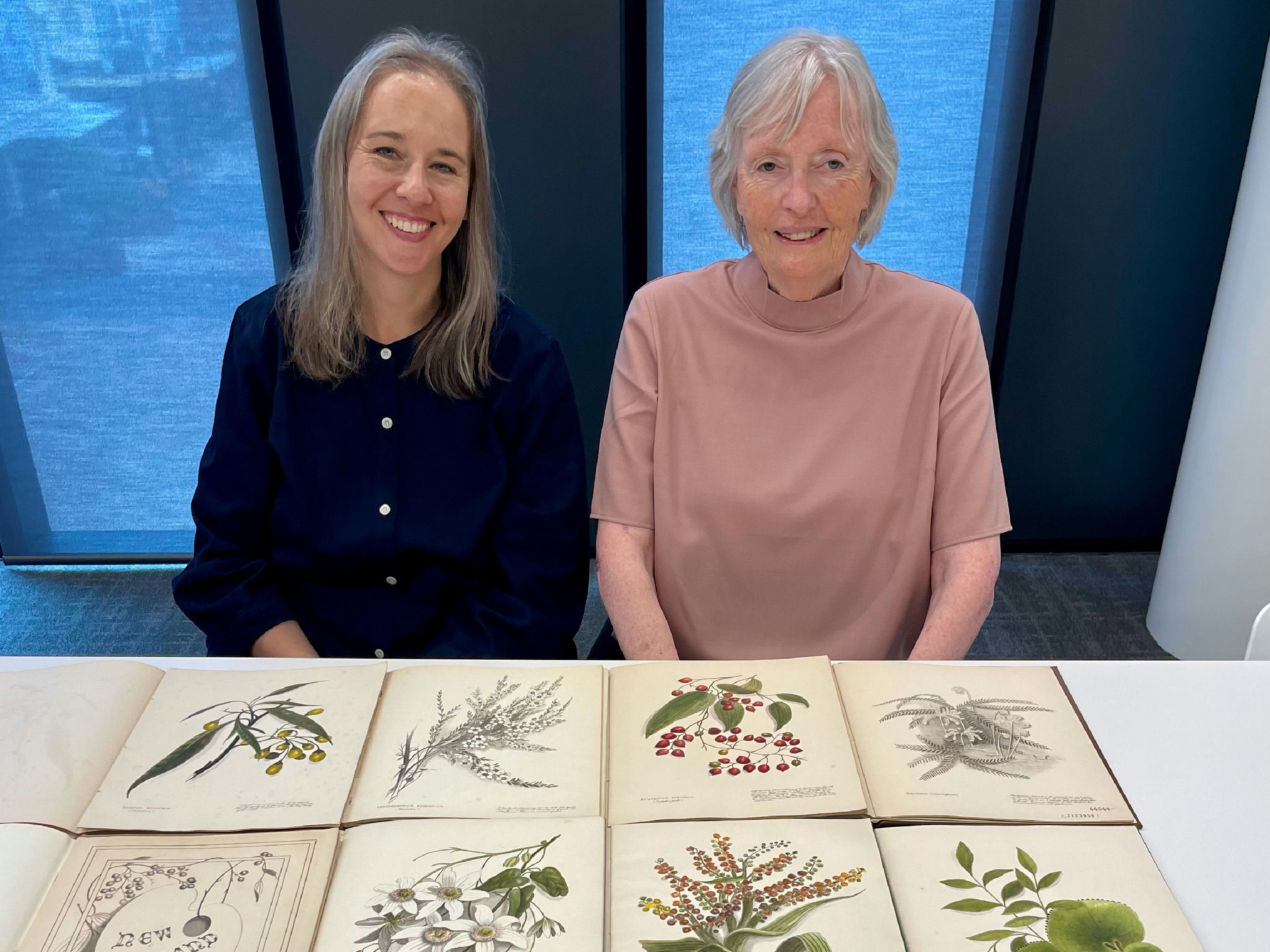 Two women sit at a table with botanical illustrations of native new zealand plants spread out..